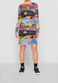 Persona vestida con un vestido colorido adornado con caras sonrientes y patrones abstractos, combinado con calcetas negras y zapatillas con un estampado de caras sonrientes.