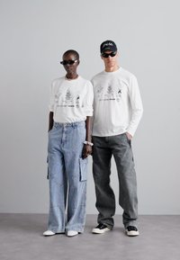 Chemises blanches à manches longues avec des motifs, associées à des pantalons cargo en denim bleu et à des pantalons gris. Les accessoires incluent des lunettes de soleil et une casquette.