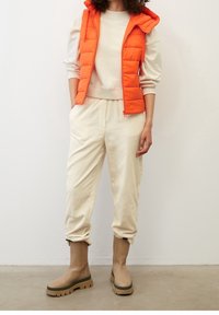 Orangene Puffweste mit Kapuze über einem beigen Sweatshirt, kombiniert mit cremefarbenen Cordhosen und sandfarbenen wasserfesten Stiefeln mit grünen Sohlen.