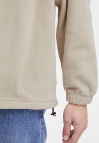 Beige fleece pullover with ribbed cuffs, elastic hem, and adjustable drawstring. Soft texture and casual design. Features a side tag.