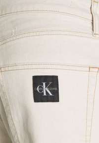 Krémové denimové kalhoty s viditelným prošíváním, se čtvercovou černou etiketou, na které je bílé logo s nápisy "CK" a "Calvin Klein Jeans."
