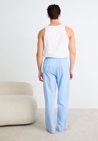 Helle blaue, leichte Baumwollschlafanzughosen mit einem elastischen Bund, kombiniert mit einem weißen ärmellosen Top, stehend auf einem weichen beige Teppich.