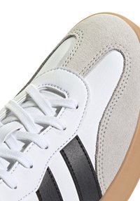 Baskets blanches avec des bandes en daim gris et cuir noir, dotées de lacets blancs, d'une tige texturée et d'une semelle en caoutchouc gomme.