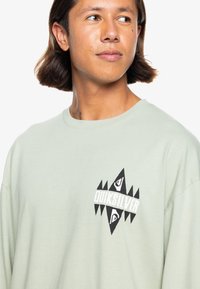 Long-sleeve T-shirt in light green cotton with a black geometric logo on the chest, featuring a round neck and relaxed fit.