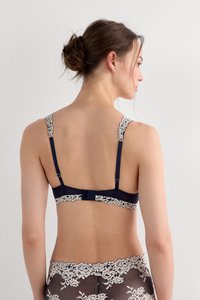 Žena zobrazená zezadu, oblečená v tmavě modré podprsence a odpovídajících krajkových kalhotkách s béžovými květinovými vzory, vlasy svázané do úhledného drdolu.