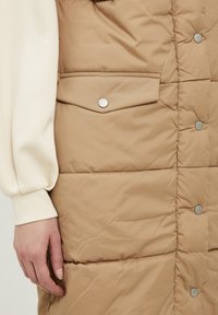 Beige gewatteerd vest met zilverkleurige drukknoopsluitingen, voorzien van een borstzak en een doorgestikte textuur. Crèmekleurig shirt met lange mouwen gedeeltelijk zichtbaar.