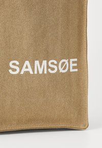 Braune Stofftragetasche mit fettem weißen Text "SAM SøE", mit strukturierter Oberfläche und genähten Kanten. Einfaches rechteckiges Design.