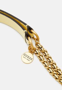 Żółto-czarny skórzany pasek z złotym metalowym łańcuszkiem. Posiada okrągłą złotą plakietkę z wygrawerowanym napisem "GUESS EST.1981".