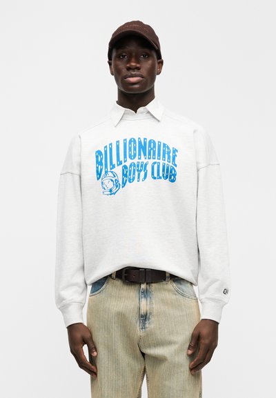 Γκρι φούτερ με μπλε γραφικό "Billionaire Boys Club" και σχέδιο αστροναύτη. Φορεμένο με ξεβαμμένο τζιν παντελόνι και καφέ καπέλο.