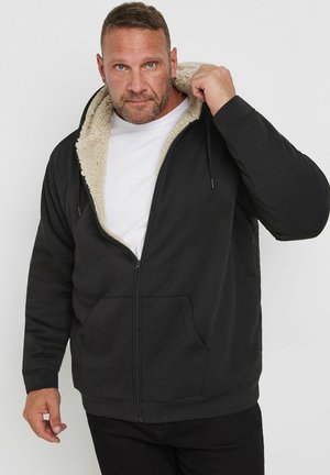 Hombre vistiendo sudadera con capucha negra con cremallera y forro de forro polar beige, sosteniendo la capucha con la mano derecha, sobre un fondo claro y liso.