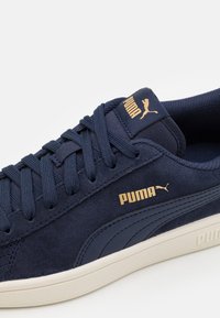 Marinblå mockasneakers med en strukturerad yta, utrustade med platta snören, en guldfärgad Puma-logotyp och en lätt, krämfärgad sula.