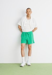 Pantaloni scurți verzi, cămașă albă perforată, top alb și adidași albi cu accente gri. Modelul stă pe o suprafață verde, în fața unui perete alb.