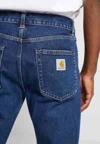 Dark blue denim jeans feature a straight leg design, visible stitching, and a square patch with a yellow logo on the back pocket.