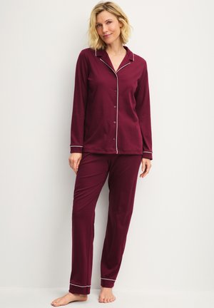 Woman wearing long-sleeve burgundy pajama set with white piping, standing barefoot against a plain light wall, smiling gently.