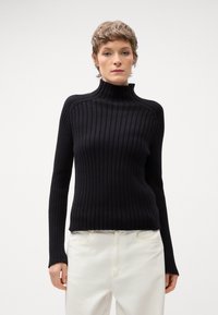 Pull à col roulé noir à côtes, à manches longues, design ajusté et tissu texturé, associé à un pantalon couleur crème.