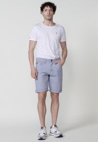 Vīrietis ar viļņainiem matiem, tērpies baltā t-kreklā, gaiši zilos sarautajos šortos un baltās čības, stāv ar vienu roku kabatā.