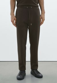 Braune Jogginghosen mit einem Kordelzugbund, schmal zulaufenden Beinen und elastischen Bündchen, kombiniert mit schwarzen Sneakers in einer minimalistischen Umgebung.
