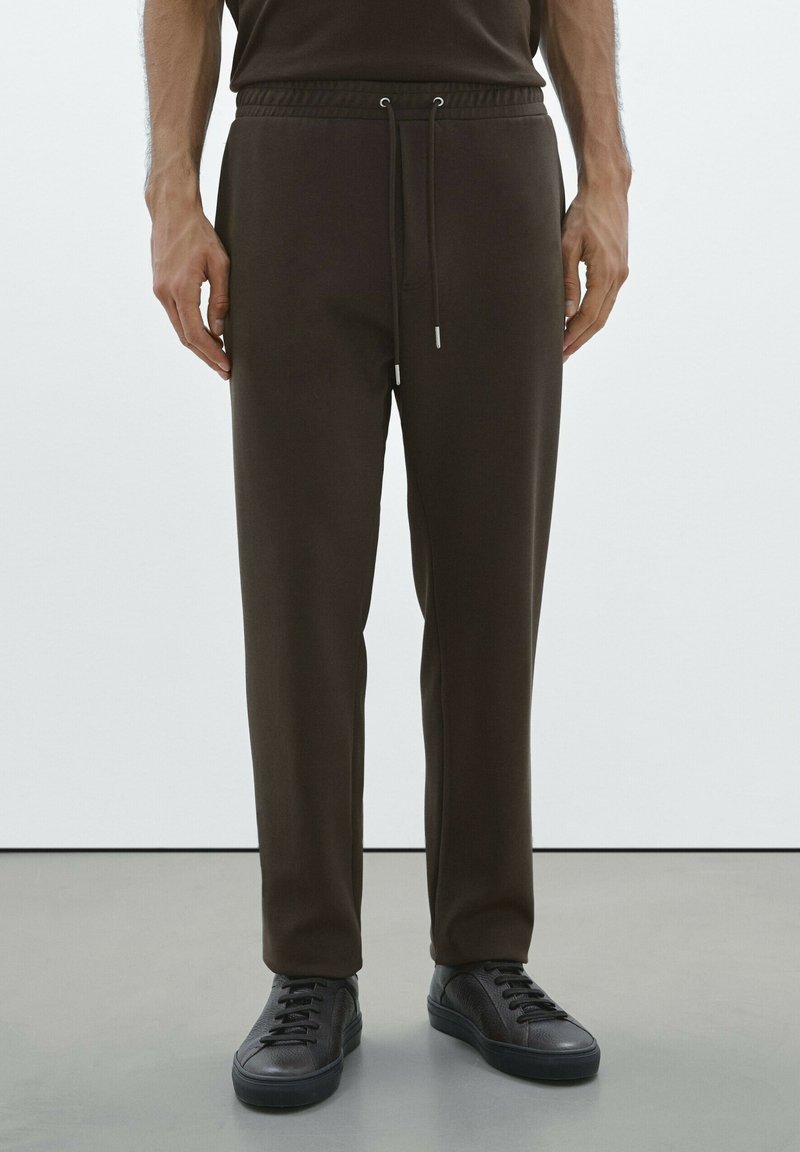 Bruine joggerbroeken met een trekkoord in de taille, taps toelopende benen en elastische boorden, gecombineerd met zwarte sneakers in een minimalistische setting.
