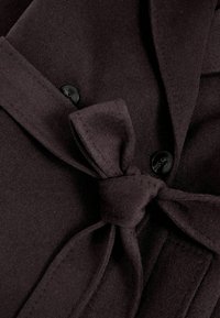 Tumši brūns vilnas mētelis ar jostu un melnu pogu aizdari, ar mīkstu tekstūru un atlocītu apkakli. Redzama smalka šuvuma detaļas.