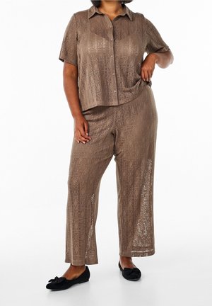 Femme portant une chemise marron à manches courtes à motifs et un pantalon large assorti, associée à des ballerines noires à nœud, debout la main sur la hanche.
