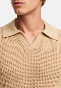 Beige strikket genser med V-hals og kontrasterende krage. Teksturen er ribbestrikket med et koselig utseende og myk stoffoverflate.