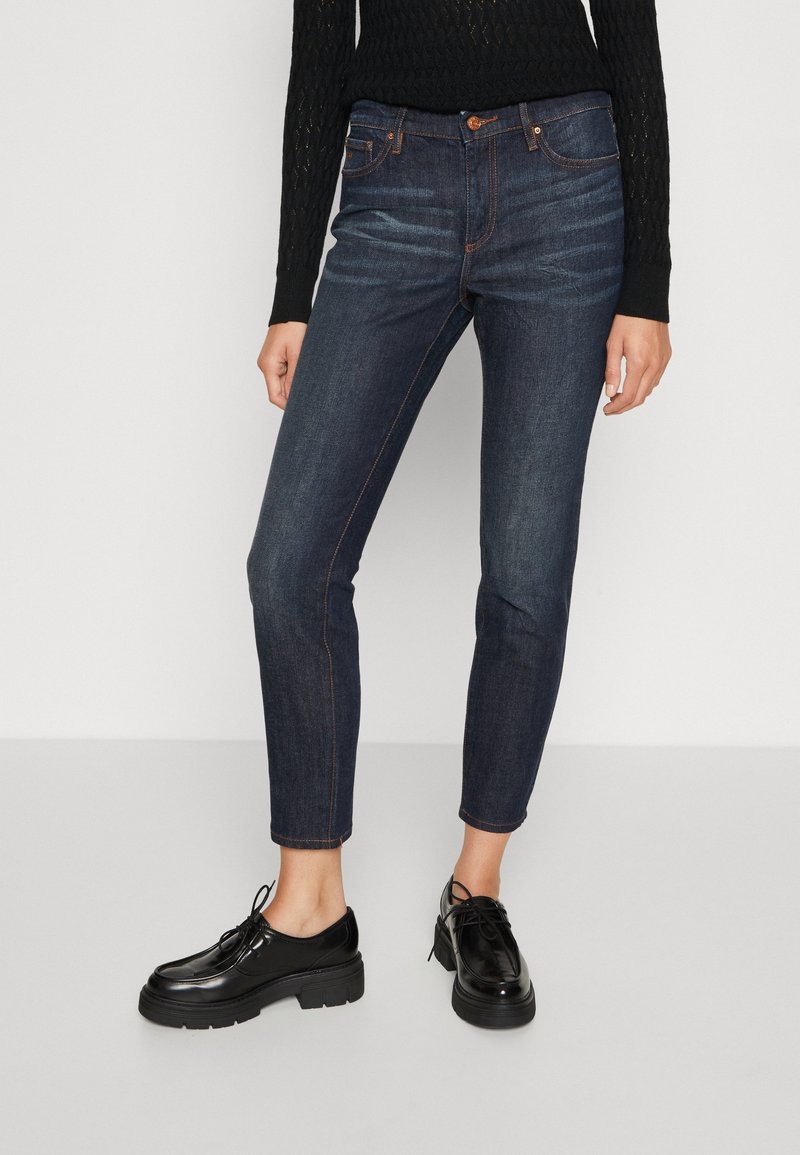 Jean en denim bleu foncé à coupe slim, avec coutures claires, fermeture par bouton et design cinq poches. Associé à des chaussures noires brillantes.