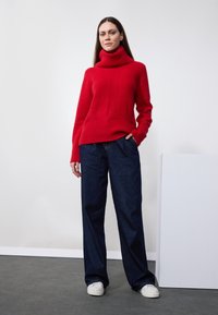 Pull à col roulé en tricot rouge avec une texture côtelée, associé à un jean en denim bleu foncé à coupe ample et des baskets blanches. Fond de mur blanc.