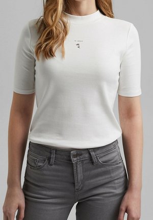 Femme portant un haut blanc à manches courtes avec un petit texte et un motif floral près du col, associé à un jean gris ajusté.