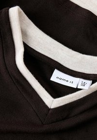 Tričko s výstřihem do V, hnědé barvy s krémovým výstřihem. Obsahuje textilní štítek s nápisem "name it" a podrobnostmi o velikosti. Jemná textura.