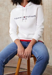 Hvid hættetrøje med snore og "TOMMY HILFIGER"-logo. Kombineret med blå jeans. Accentlogo på ærmet. Sidder på en træskammel.