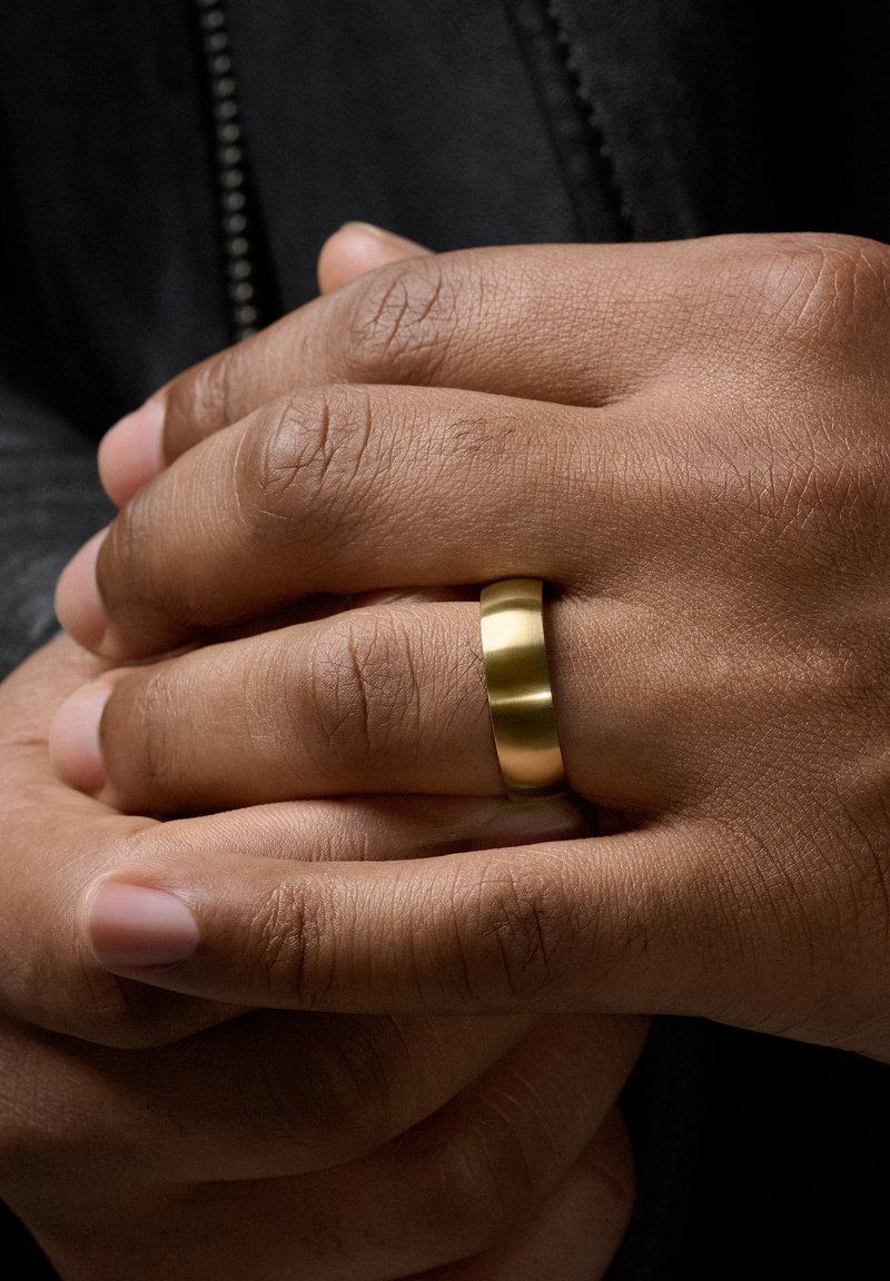 Eine goldfarbene Metallring mit einer glatten, runden Form, getragen an einer Hand mit mittlerem Hautton, umgeben von texturierter Haut.