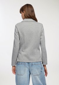 Giacca grigia attillata con colletto a risvolto e maniche lunghe. Texture liscia, design strutturato, abbinata a jeans a gamba larga blu chiaro.