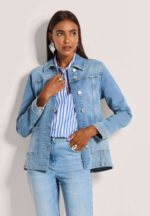 Frau trägt hellblaue Jeansjacke und Jeans mit blau-weiß gestreiftem Hemd, posiert mit einer Hand in der Tasche und der anderen nahe am Kragen.