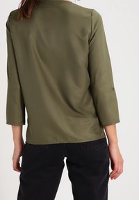 Donna che indossa una blusa a maniche lunghe verde oliva e pantaloni neri, vista da dietro su uno sfondo uniforme.