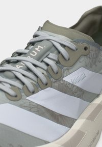 Gaiši pelēks sporta apavu ar baltām svītrām, sarežģītu ziedu rakstu, pelēkām šņorēm un tekstu "A-R-U-M" uz mēles.