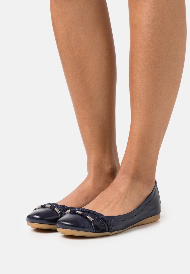 Ballerines en cuir bleu marine avec des détails tressés et des perles métalliques, portées pieds nus sur un fond uni.