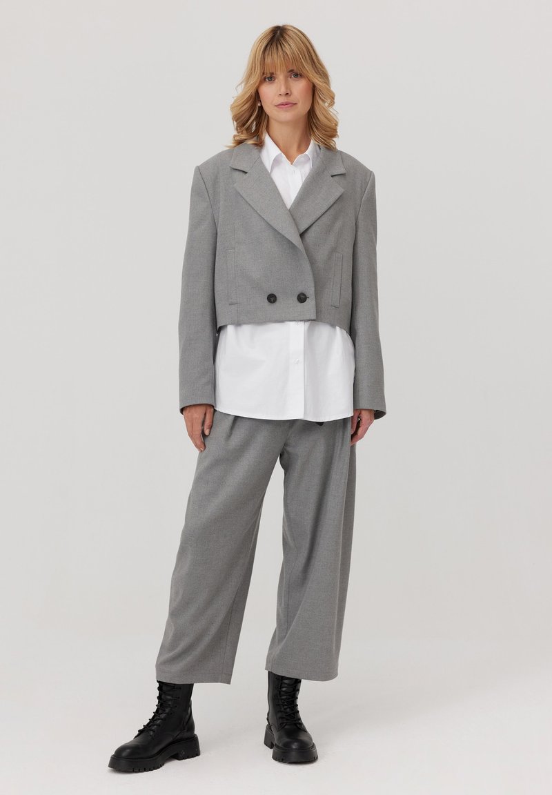 Szary krótki blezer z podwójnymi guzikami, zestawiony z białą koszulą i szerokimi szarymi spodniami, noszony z czarnymi sznurowanymi botkami. Gładka faktura.