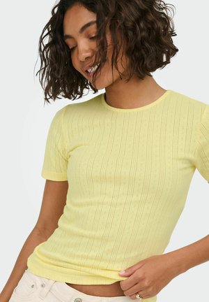 Young woman with curly hair wearing a pale yellow short-sleeve knit top, smiling and looking down with one hand holding the hem.