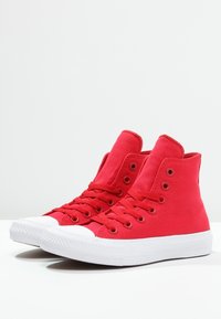 Sneakers alti rossi in tela con suola piatta in gomma bianca, punta rotonda e sei occhielli per i lacci su ciascun lato.