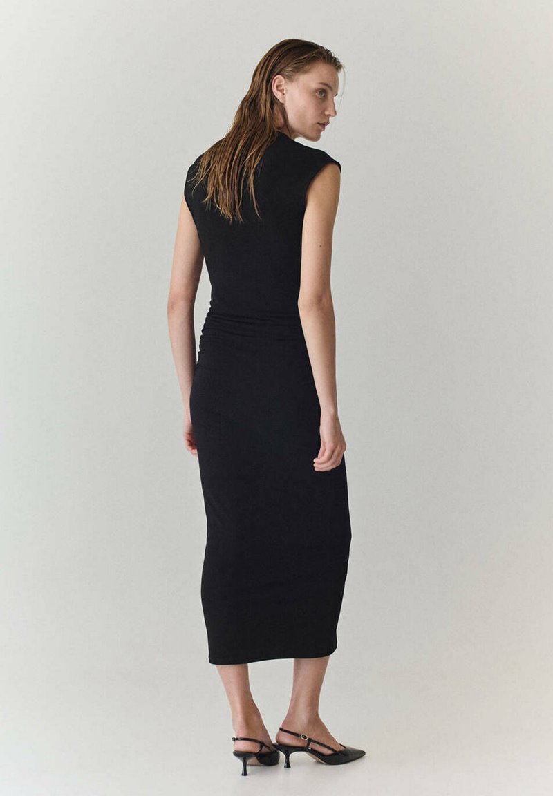 Femme aux cheveux mouillés portant une robe midi noire sans manches et des chaussures slingback noires à talons bas, debout devant un fond uni.