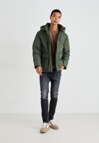 Polstret grønn jakke med hette, to frontlommer og glidelås. Bæres over en brun skjorte og mørke jeans, komplementert med lyse sneakers.