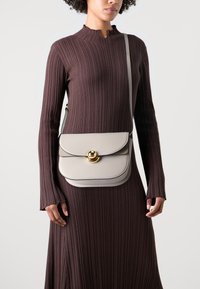 Borsa in pelle grigia con patta arrotondata, dettagli in metallo dorato e tracolla regolabile, portata da una persona con un vestito marrone texturizzato.