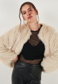 Faux fur jas in lichtbeige met een fluffy textuur, wijde mouwen en een open voorkant, gedragen over een zwarte doorschijnende top en leren broek.