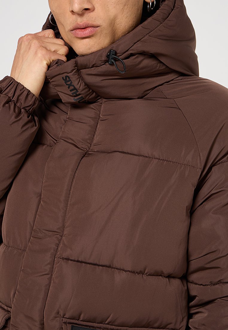 Braune Pufferjacke mit Kapuze, aus strukturiertem Stoff und mit elastischen Bündchen. Mit Kordelzugverschluss und einem Logo auf der Brust.