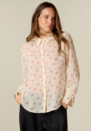 Frau mit langen Haaren, die eine hellcremefarbene Bluse mit pinkem Blumenmuster, langen Ärmeln und schwarzen Hosen trägt, Hände in den Taschen.