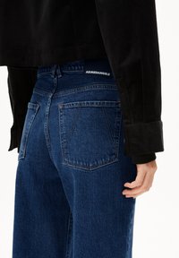Donkerblauwe denim high-waisted jeans met twee achterzakken, lichte stiksel en een zichtbaar label op de tailleband. Een gedeeltelijk zichtbare corduroy jas.
