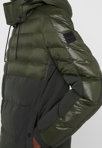 Mørkegrøn polstret jakke med hætte, sort lynlås, knappede lommer og et sort Boss-logo patch på den øverste ærme.