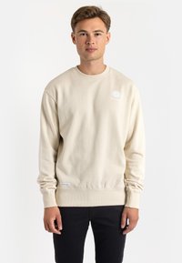 Beige sweatshirt with long sleeves, round neck, and subtle white logo on the chest. Smooth cotton texture, ribbed cuffs, and hem.