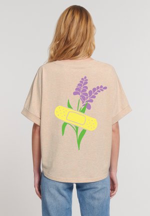 A woman with long hair wears a casual beige t-shirt featuring a yellow bandage and purple flowers printed on the back.