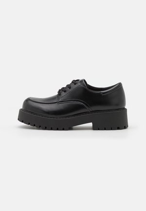 Zwarte leren veterboot met dikke profielzool, ronde neus en lage blokhak op een witte achtergrond.